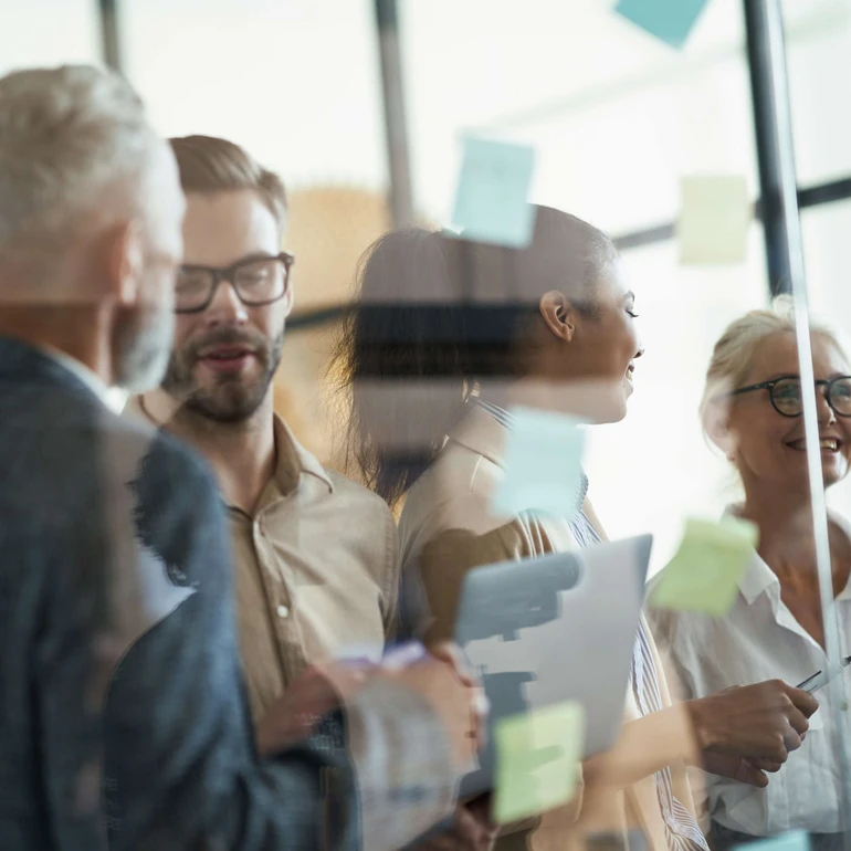 Teamwork im kreativen Austausch Mehrere Kolleginnen und Kollegen stehen vor Glaswänden mit Haftnotizen und besprechen Ideen.