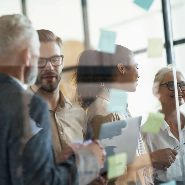 Teamwork im kreativen Austausch Mehrere Kolleginnen und Kollegen stehen vor Glaswänden mit Haftnotizen und besprechen Ideen.