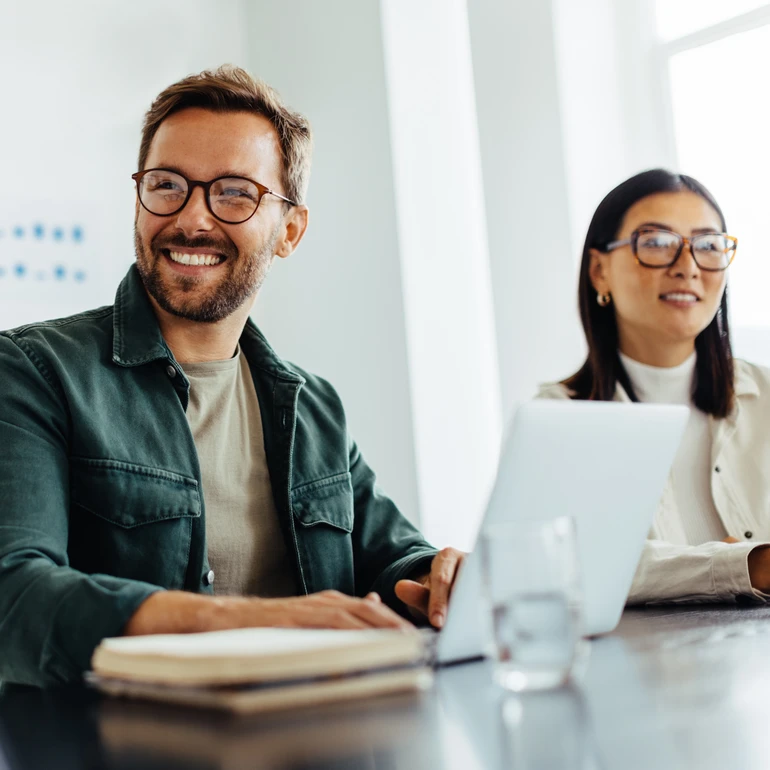 Teammeeting im Büro Mann mit offenem grünen Hemd, Brille und Bart sowie eine Frau mit weißem Blazer und Brille lachen gemeinsam vor einem Laptop in einem Meeting
