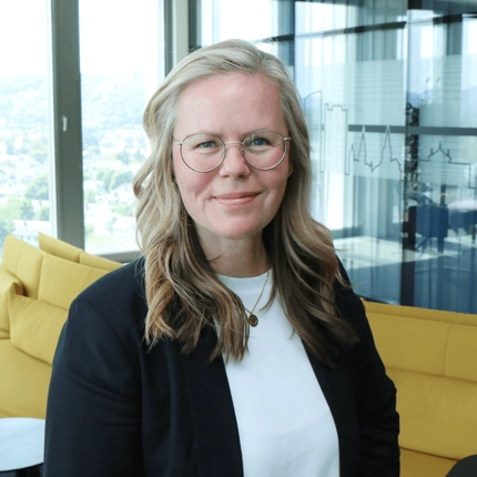 CONET Ansprechpartnerin für Karriere – Porträtfoto Porträt einer lächelnden Frau mit Brille und langen, blond-braunen Haaren, die in einem modernen Büro vor einer gelben Couch steht.