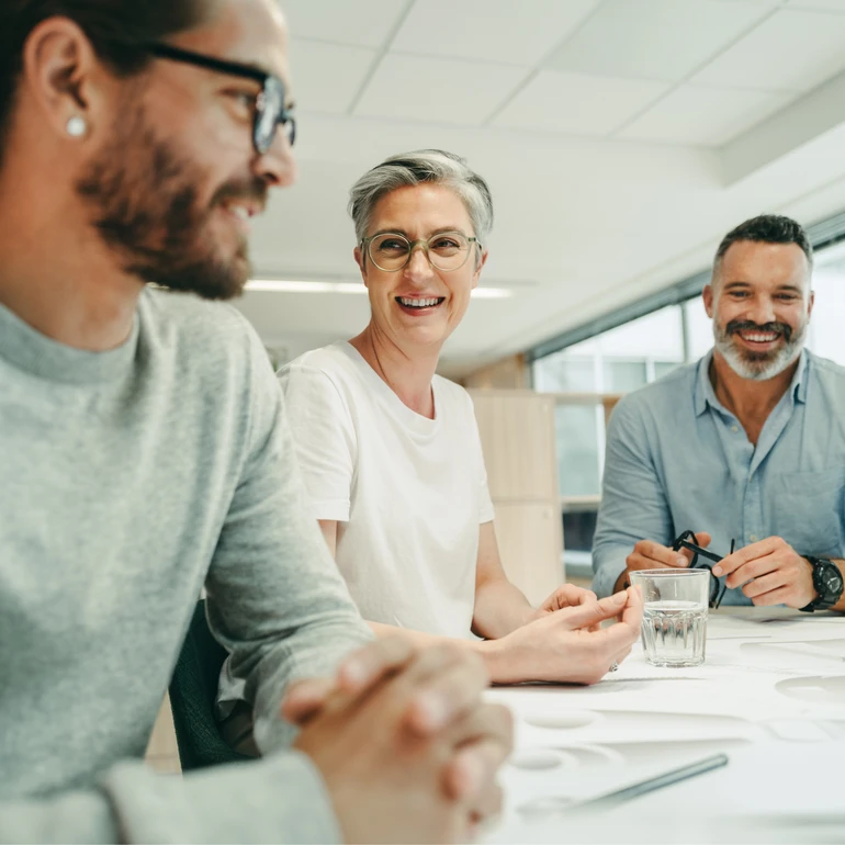 Teammeeting im Büro Drei Personen sitzen lachend an einem Besprechungstisch, ein junger Mann mit Bart im grauen Shirt, eine Frau mit kurzen grauen Haaren und ein Mann mit Bart im Hintergrund
