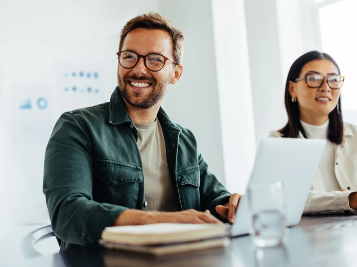 Teammeeting im Büro Mann mit offenem grünen Hemd, Brille und Bart sowie eine Frau mit weißem Blazer und Brille lachen gemeinsam vor einem Laptop in einem Meeting