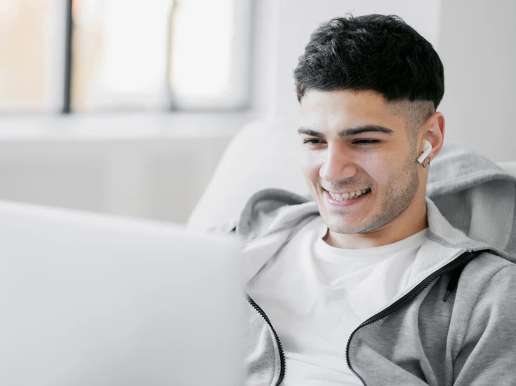 Person am Laptop Junger Mann mit Airpods in hellgrauer Jacke lächelt beim Arbeiten am Laptop