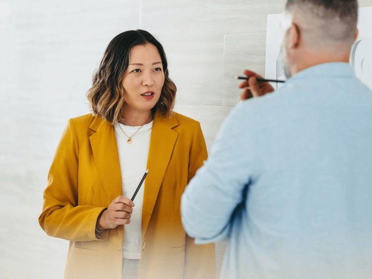 Professionals & Experts Frau in gelbem Blazer spricht mit einer Person und hält einen Stift in der Hand