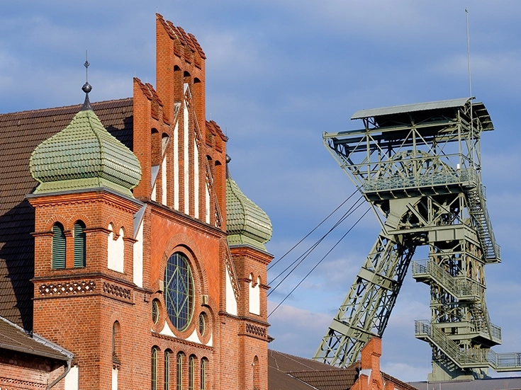 Backsteinfassade eines historischen Gebäudes der Zeche Zollern in Dortmund mit grünlich verzierten Turmkuppeln und großer Rundbogenfensterfront; daneben ragt ein stählerner Förderturm vor blauem Himmel auf.