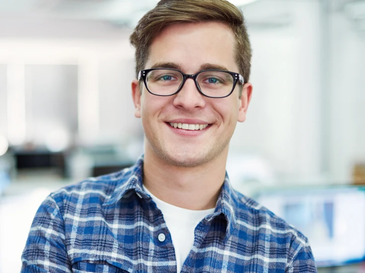 Portrait im Büro Junger Mann mit Brille im karierten Hemd lächelt in die Kamera, heller Büroraum im Hintergrund
