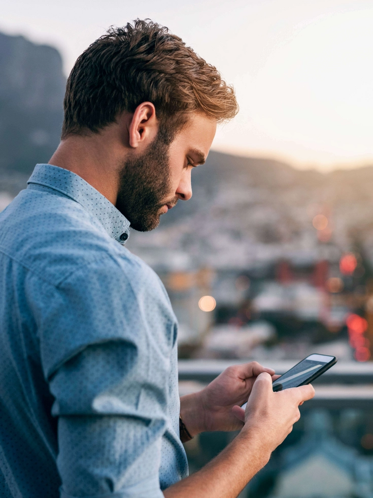 Mobiler Arbeitsplatz mit urbanem Ausblick Mann steht auf einer Dachterrasse und nutzt sein Smartphone mit Blick über eine Stadtlandschaft.