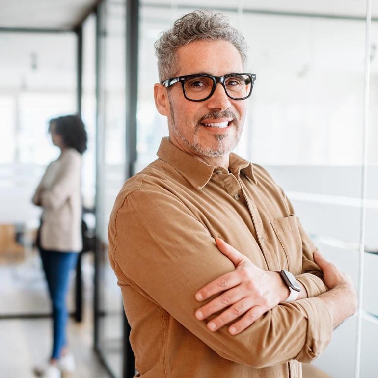 Portrait im Büro Mann mit grauen Haaren und Brille im braunen Hemd steht mit verschränkten Armen und lächelt in die Kamera, Büroraum im Hintergrund